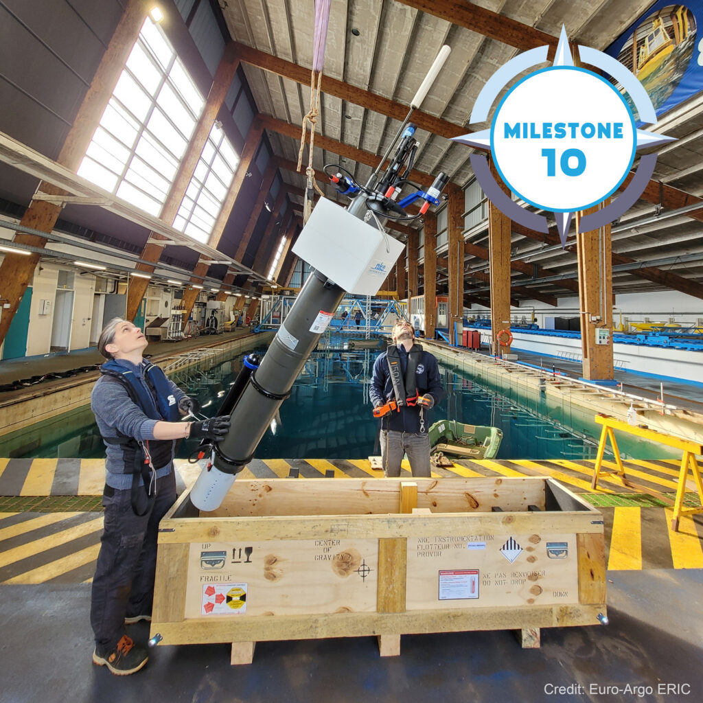 The setting of this picture is a laboratory in France, which looks like a large indoor swimming pool with wooden constructions. Two scientists in the front are looking up at a pulley system pulling up a Biogeochemical Argo float from a wooden box. Behind them is a water pool. On the top right corner of the picture, a TRICUSO branded milestone badge has been placed, showing 'milestone 10'.