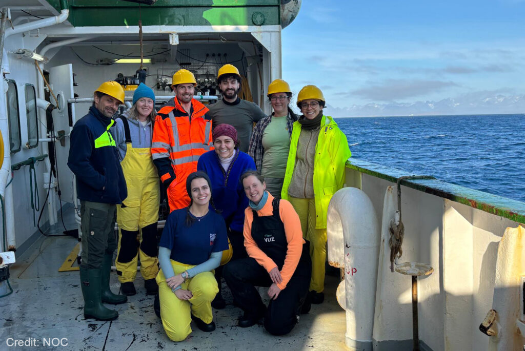 A group picture of nine people on board of a ship. Most of them wearing yellow helmets and other fluorescent work clothing. A blue sky and ocean in the background.