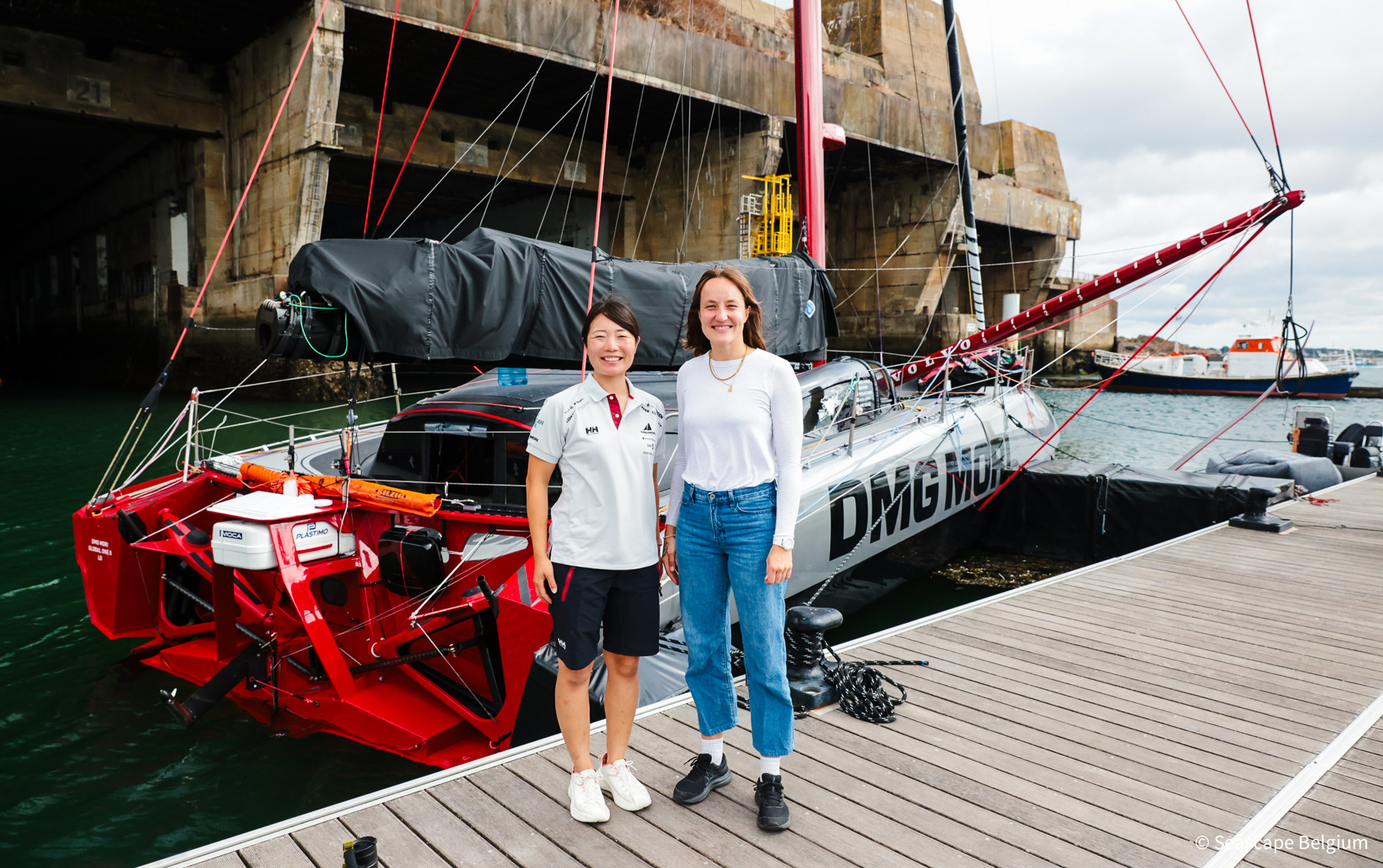 Two women, a sailor and a scientist, posing in front of the DMG MORI Global One Racing yacht in the harbour.