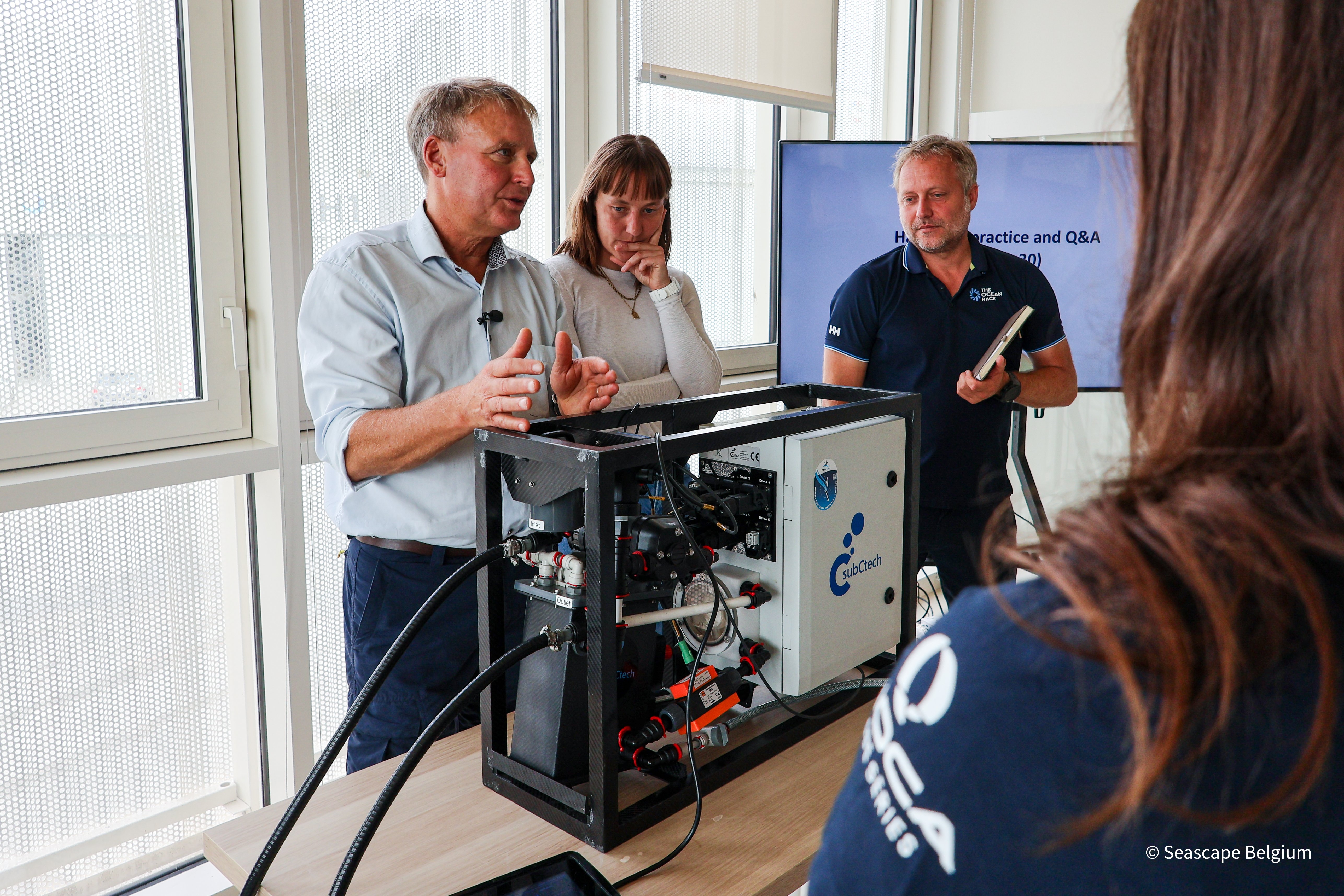 Close-up of two scientists giving a hands-on presentation about the OceanPack instrument and one sailor listening attentively.