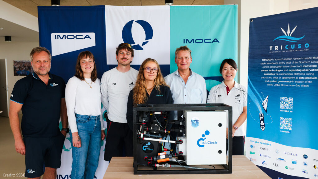 A group picture of three men and three women smiling at the camera and posing behind the SubCtech OceanPack instrument that will be installed on the DMG MORI Global One racing yacht. In the background are the IMOCA logo and the TRICUSO banner.